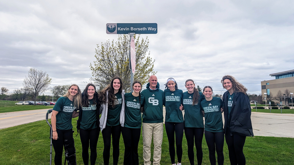 Kevin Borseth stands with members of the Green Bay Phoenix women's basketball team under the new 'Kevin Borseth Way' street sign on campus. From left to right: Callie Genke, Bailey Butler, Jenna Guyer, Maren Westin, Borseth, Jasmine Kondrakiewicz, Sohpie Lahti, Natalie McNeal and Megan Shultz.